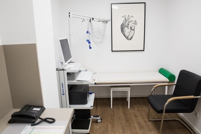Zimmer zur EKG-Untersuchung mit modernem EKG-Gerät zur kardiologischen Untersuchung, im Hintergrund anatomisches Bild eines Herzens, Untersuchungsliege für EKG- und Blutdruckmessungen und Infusionstherapie